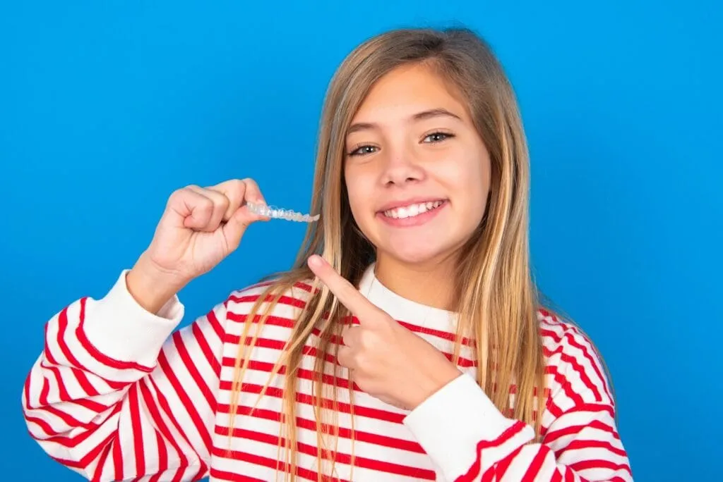 Girl wearing a striped sweater holding a clear aligner and smiling, showcasing confidence in orthodontic treatment options like Invisalign for kids.