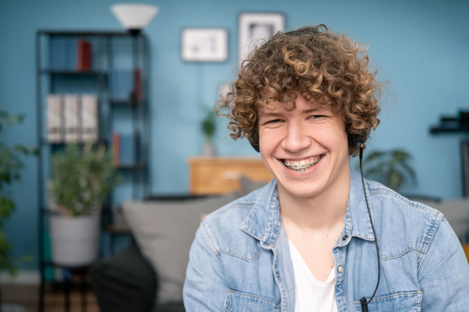 Smiling young man with curly hair and braces listening to music in a cozy living room, emphasizing the importance of orthodontic care and maintaining a healthy smile.